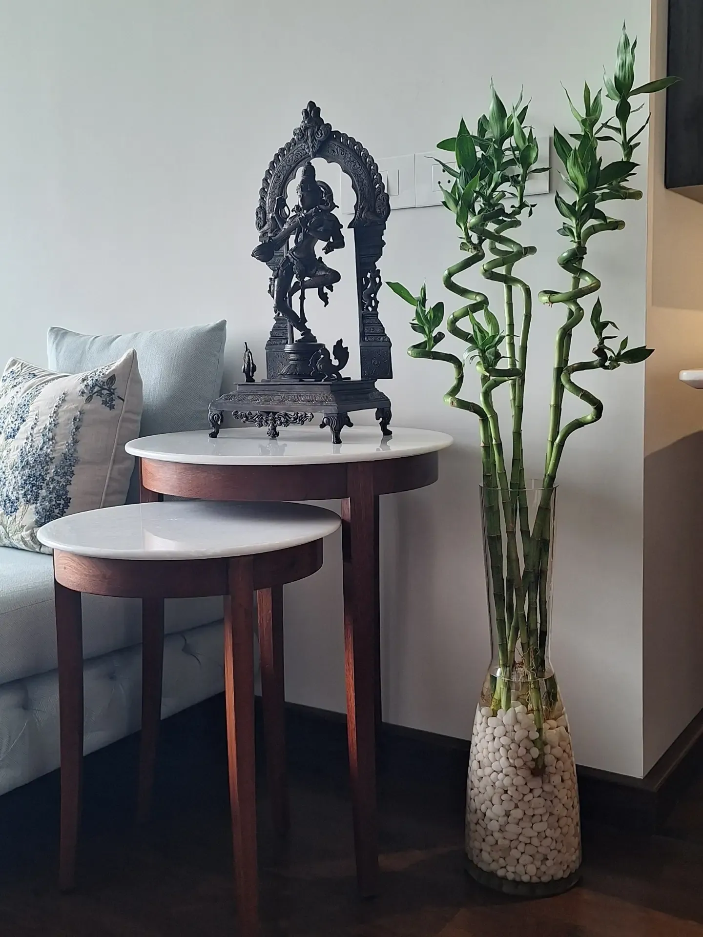 Lucky bamboo in a tall glass vase of white pebbles beside nesting marble side tables and a bronze idol, in a home in Oshiwara, Andheri West, Mumbai