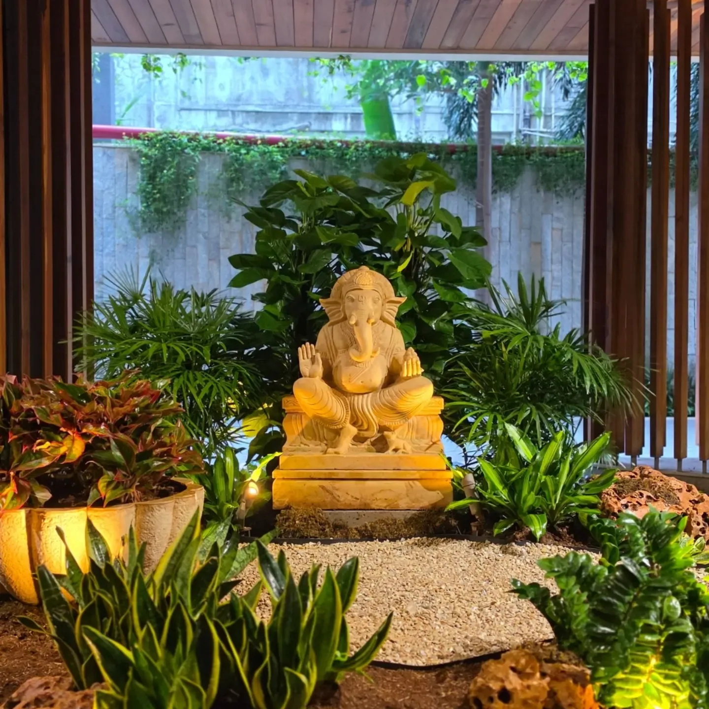 Sandstone Ganpati idol framed by layered plants including palms, aglaonema, dracaena, snake plant and ZZ plant, with gravel, laterite rocks and warm uplighting, in a residential society on Carmichael Road, Mumbai